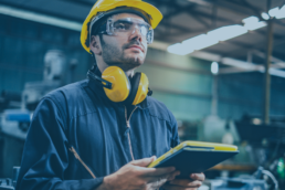 Operatore con casco, occhiali protettivi e cuffie antirumore in un impianto industriale, mentre utilizza un tablet per il monitoraggio della sicurezza con RTLS nelle aree produttive .